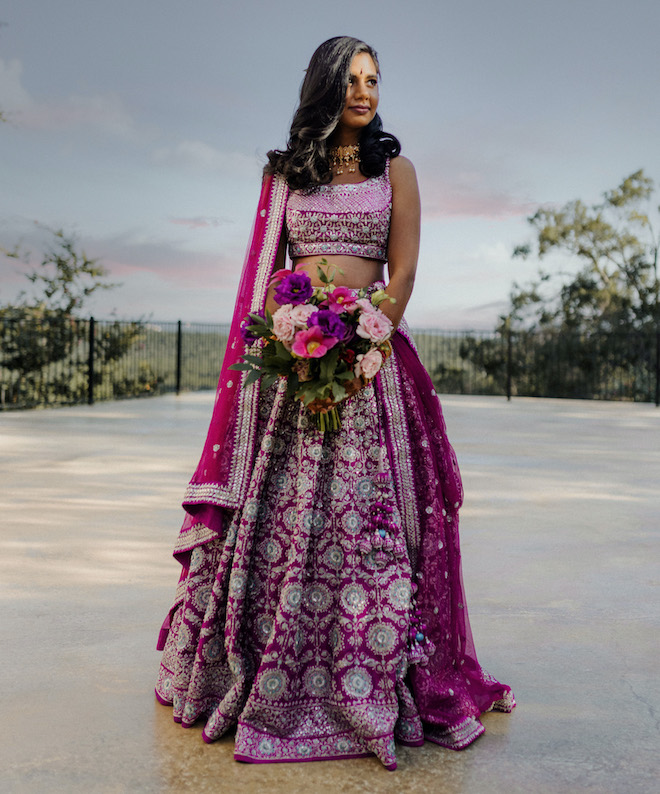 The bride stands outside the wedding venue wearing mauve colored traditional Indian wedding attire.