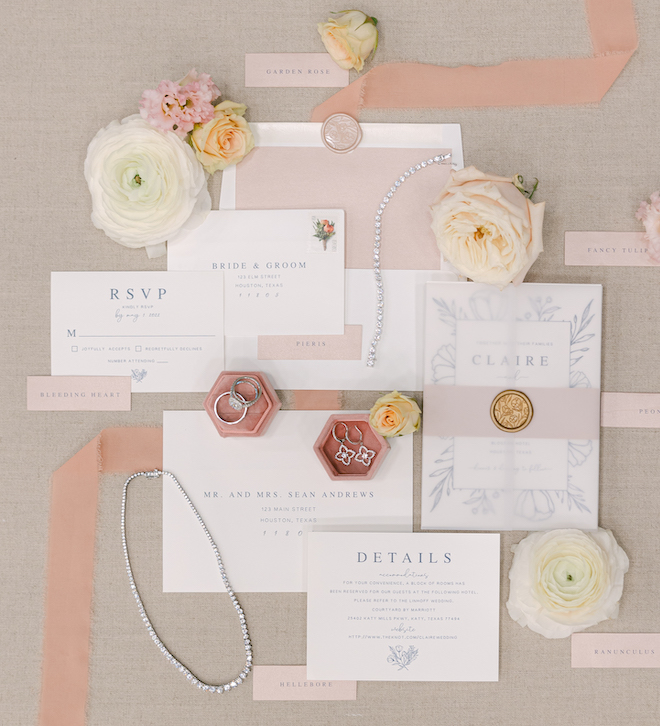 A white invitation suite with blue lettering decorated with pink accents, florals and bridal jewelry.