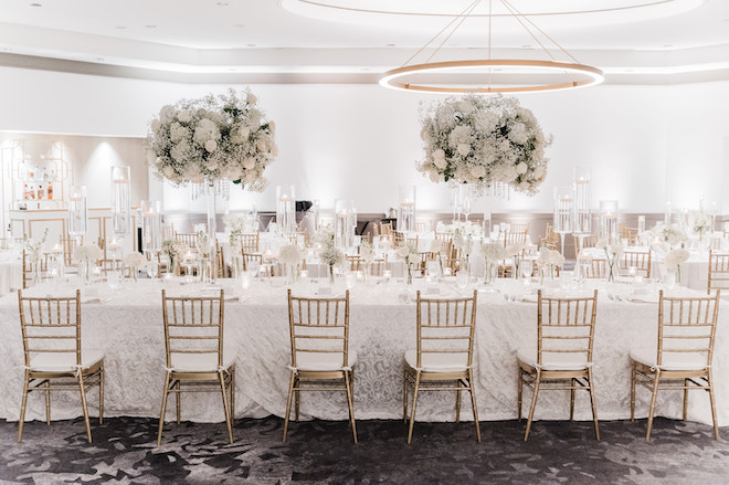 Long reception table with gold chairs, white linens and large white floral centerpieces. 