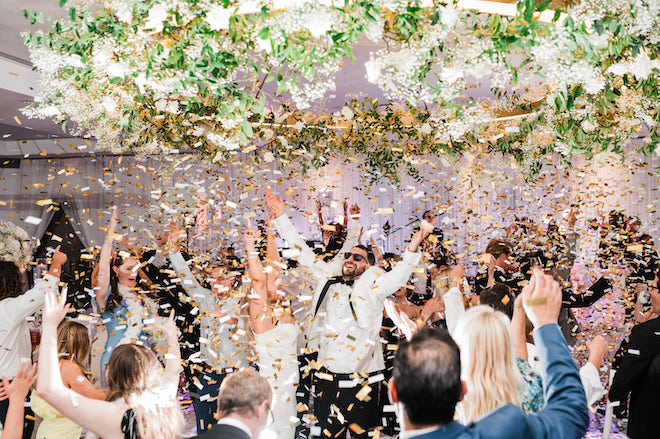 The bride and groom cheering surrounded by their guests while gold confetti falls from the ceiling. 