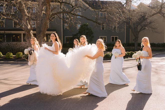 The bridesmaids hold the bride's wedding dress train as they walk to the outside ceremony.