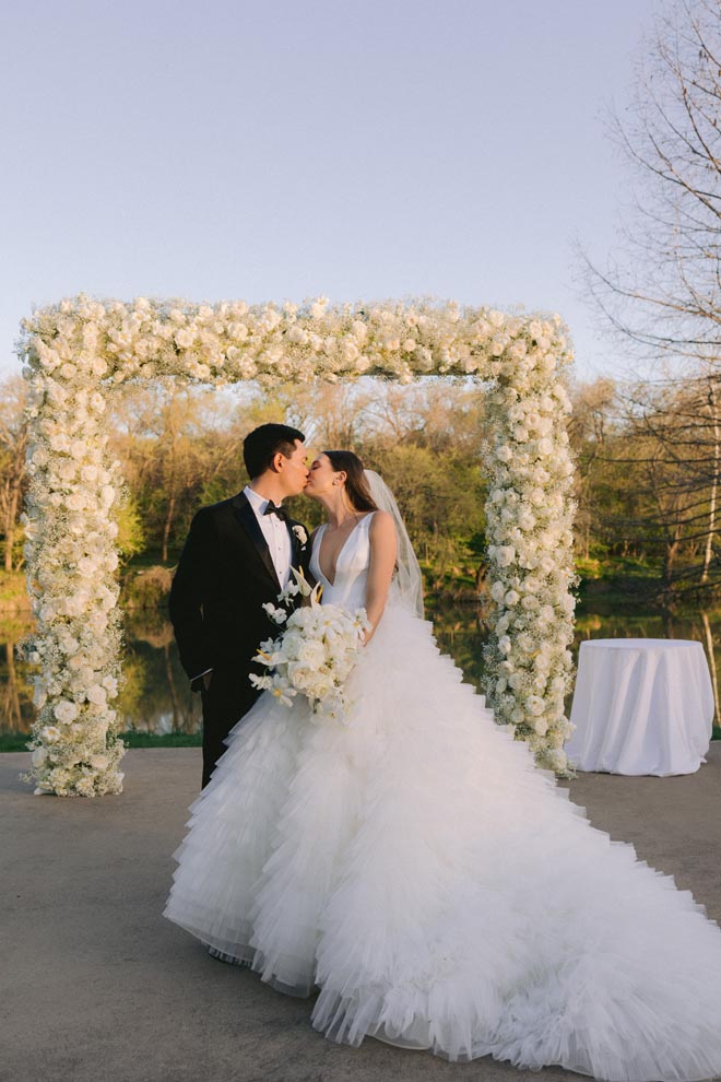 The bride and groom share a kiss under a white floral instillation at their sun-kissed al fresco wedding.