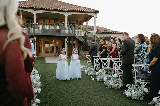 Two flower girls in white dresses walking down the baby's breath lined aisle. 