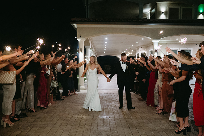 The bride and groom holding hands while the guests hold sparklers on either side of them. 