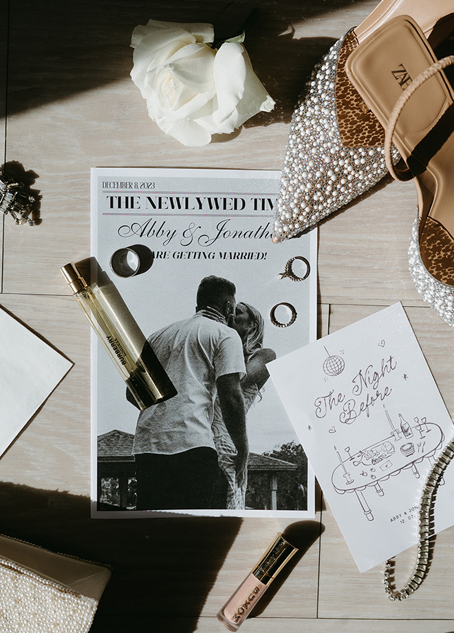 A flatlay with sparkly heels, perfume, "The Night Before" invitation and "The Newlywed Times" program. 