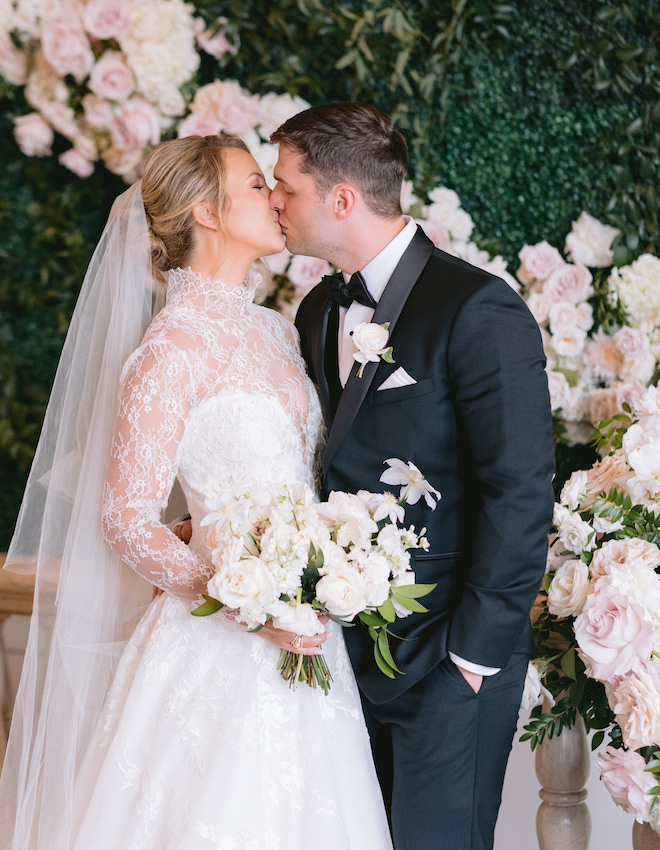 The bride and groom kissing in front of a wall of greenery with white and blush florals. 