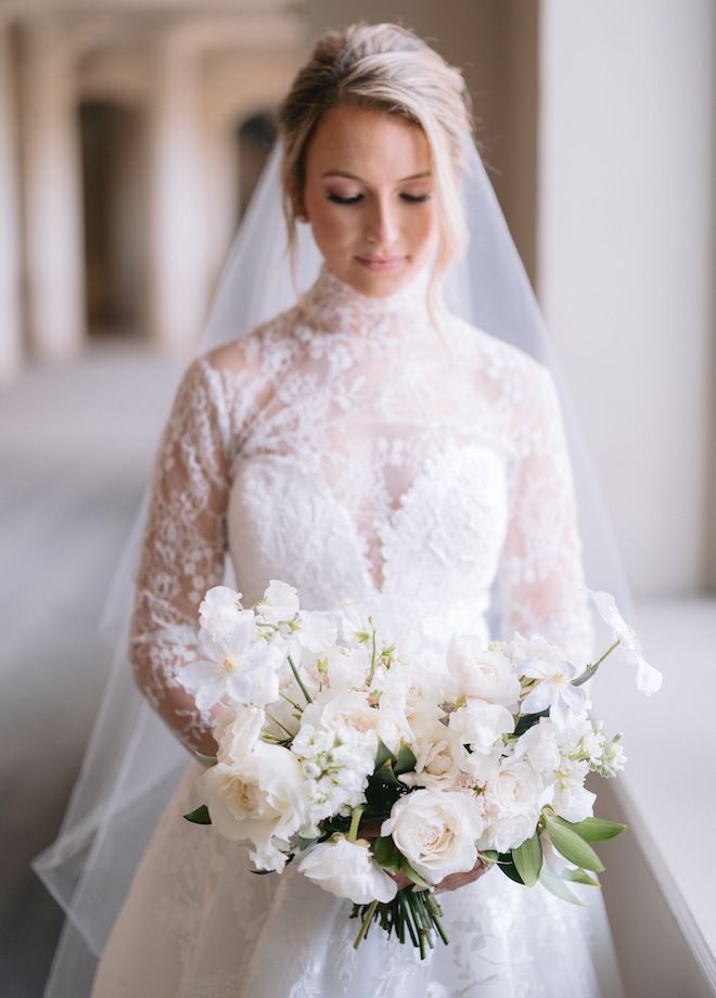 The bride wearing a long sleeve lace wedding dress looking down at her bouquet with white florals. 