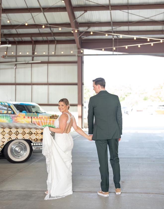 The bride and groom hold hands as they walk beside vintage cars. 