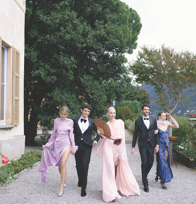 The guests walking down a road outside wearing tuxedos and bright dresses. 