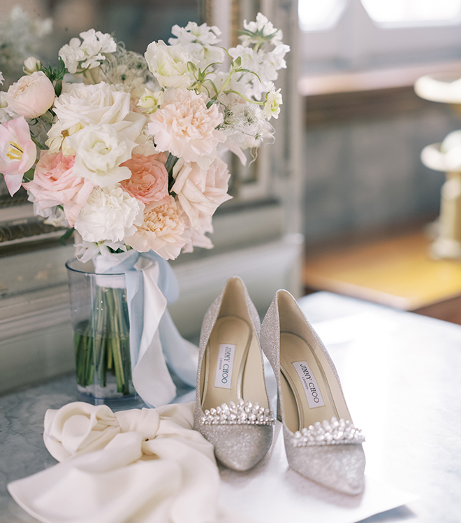 Sparkly Jimmy Choo's sitting next to a bouquet of white and pink flowers in a glass vase. 