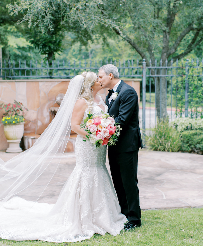 A bride and groom kissing outside while the bride holds a pink floral bouquet. 