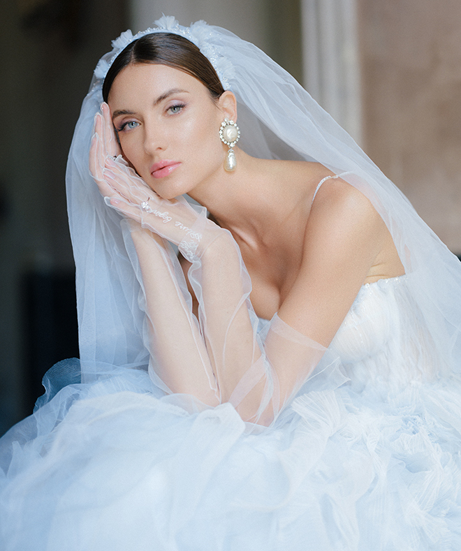 Bride wearing a blue tulle wedding dress, veil and gloves with large pearl earrings. 