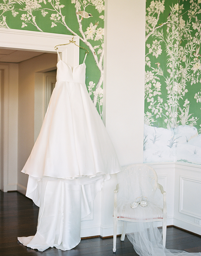 A wedding dress hanging on the door frame in a room with green and white floral wallpaper. 