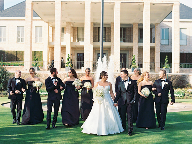 A bride, groom and their wedding party in b lack dresses and black tuxes walking outside of a country club. 