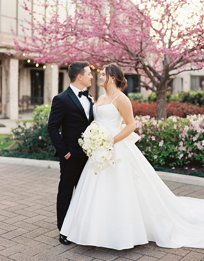 The bride and groom smiling at each other in front of a pink tree. 