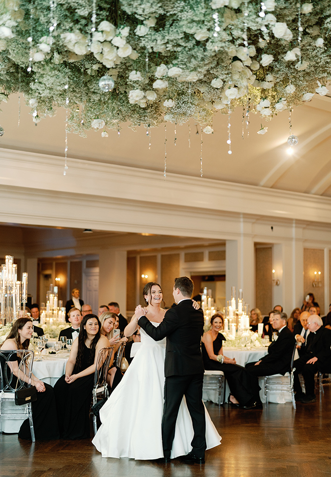 A bride and groom dancing under a hanging floral arrangement on baby's breath, white florals and crystals. 