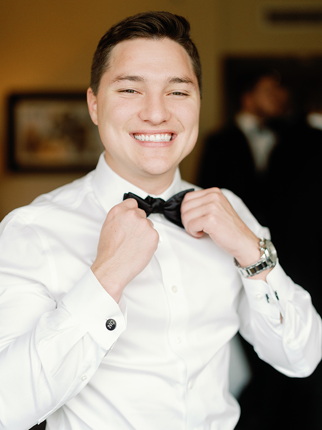 The groom smiling while adjusting his bow-tie. 