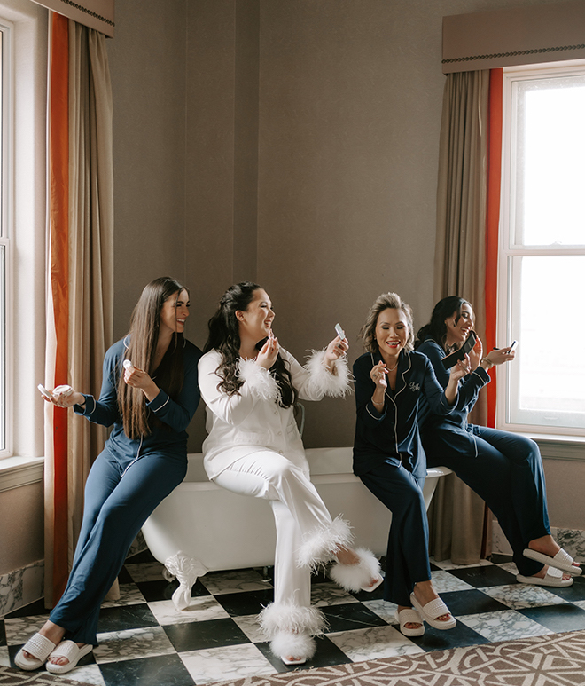 The bride and her bridesmaids in navy pajamas putting on makeup white sitting on the edge of a bath tub. 