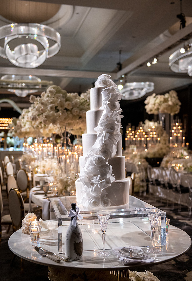 A five-tier wedding cake is placed on an acrylic cake stand for the grand ballroom reception.