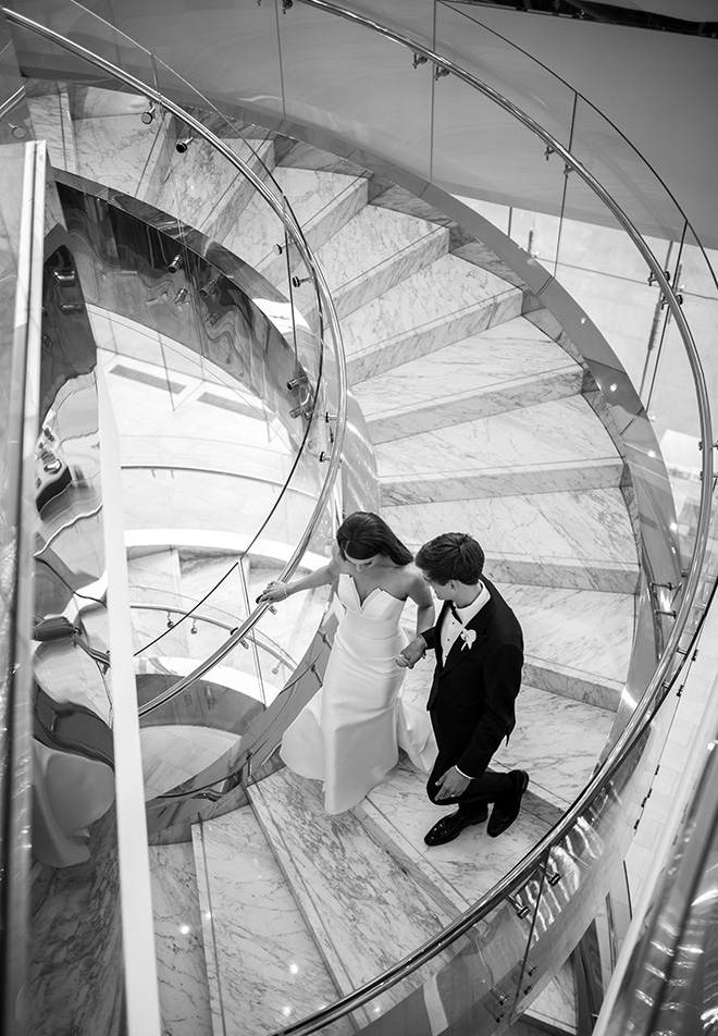 The bride and groom hold hands as they walk down the grand staircase of a Houston hotel.