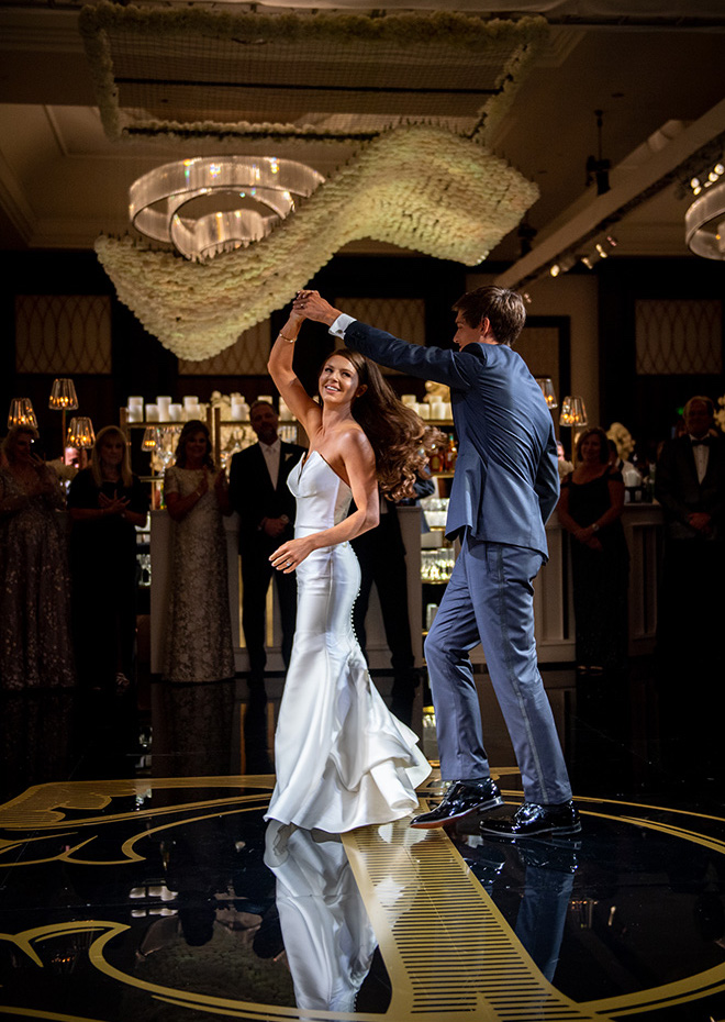 The bride and groom dance at their grand ballroom reception on a navy and gold monogrammed dance floor.