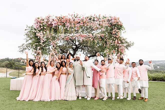 Bride, groom and the wedding party standing in front of a mandap covered in pink flowers and greenery at Omni Barton Creek Resort & Spa.