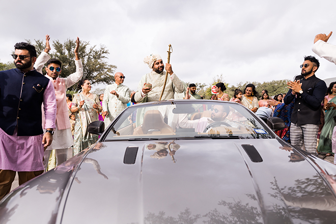 Groom in a gray sports car with guests cheering around him. 