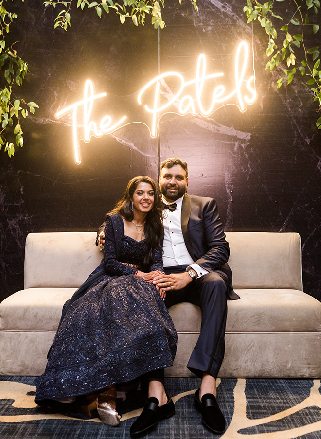 A bride and groom smiling on a couch with a lit-up sign behind them that says "The Patels"