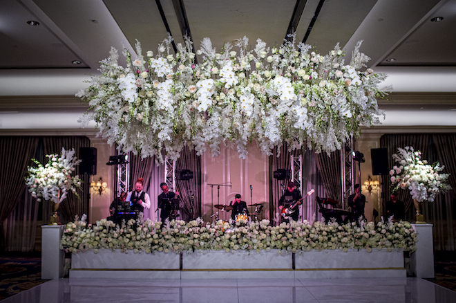 A wedding band under a canopy of white florals and greenery in front of a white dance floor. 