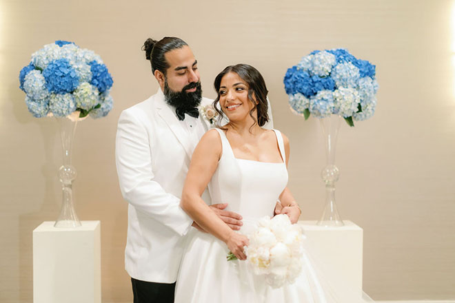 The couple stands next to blue hydrangeas floral arrangements at the ceremony in the venue's ballroom