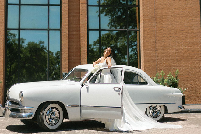 The bride stands in a vintage white car.