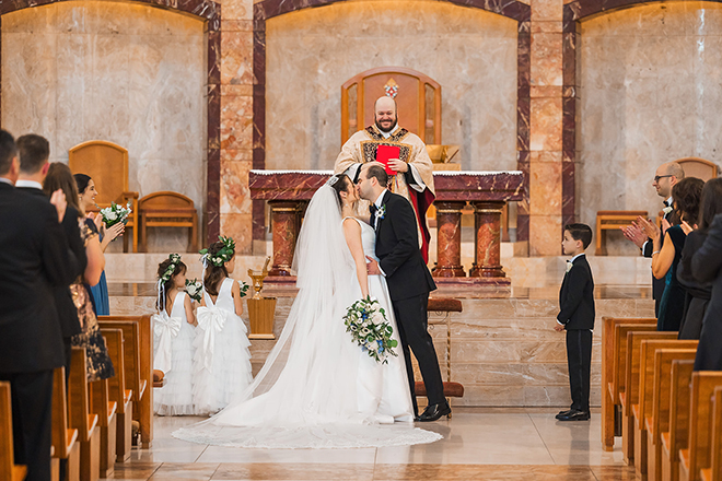 The bride and groom stand at the end of the aisle kissing as their guests rise and applaud. 