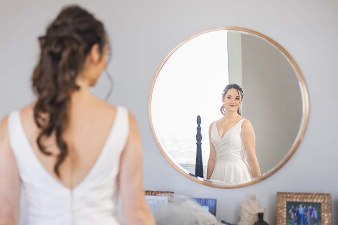 The bride smiles in the mirror at her hotel while she gets dressed in her gown. 