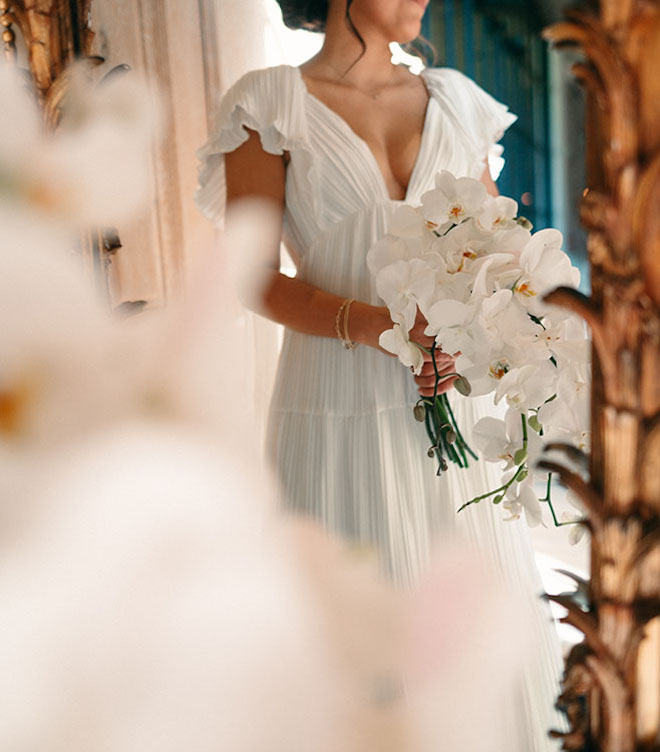 The bride stands infront of the mirror after putting her flowy dress on holding an orchid bouqet. 