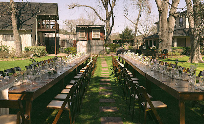 The long family-style connected reception tables fill the outside venue, trees surround the tables. 