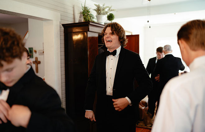 Groom smiles as he is getting ready with his groomsmen beside him. 