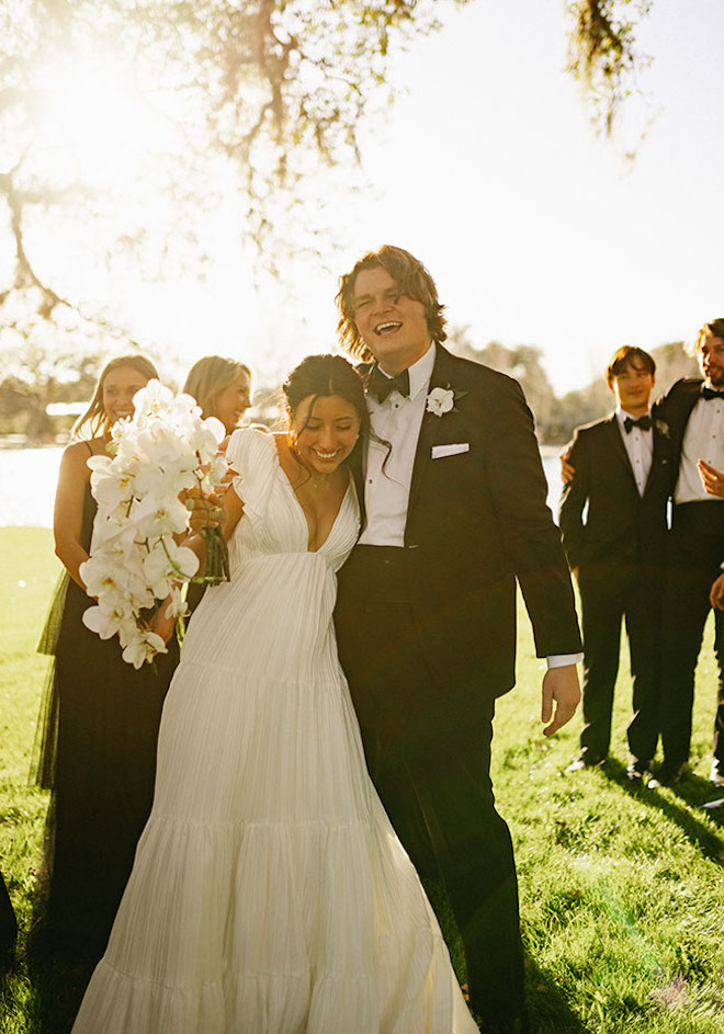 The bride and groom hug tightly smiling with their wedding party underneath a tree as the sun shines bright. 