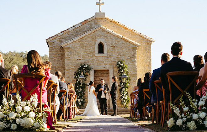 The bride and groom stand at the end of the aisle in an outside church. 