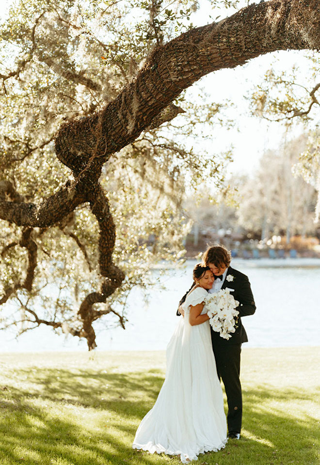 Bride and groom hug underneath an oak tree as they take their wedding photos. 