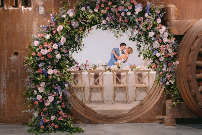 Photo of a vault and inside a couple having dinner and posing for a photo underneath florals. 