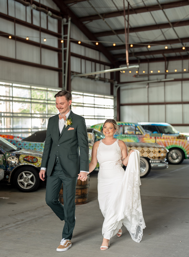 The happy couple walks off their reception smiling while bride holds her dress up with vintage cars behind them. 