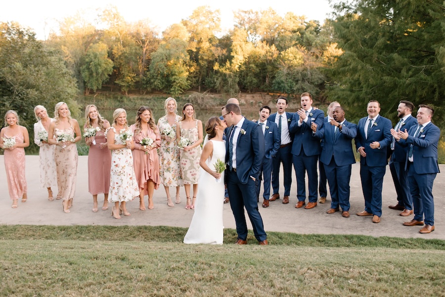 The bridal party along with the bride and groom pose for a photo and applause the couple as they kiss at their wedding ceremony. 