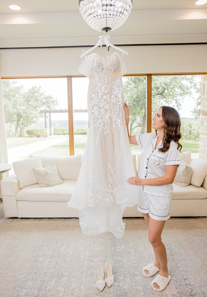 The bride stands next to her wedding dress that hangs from a chandelier. 