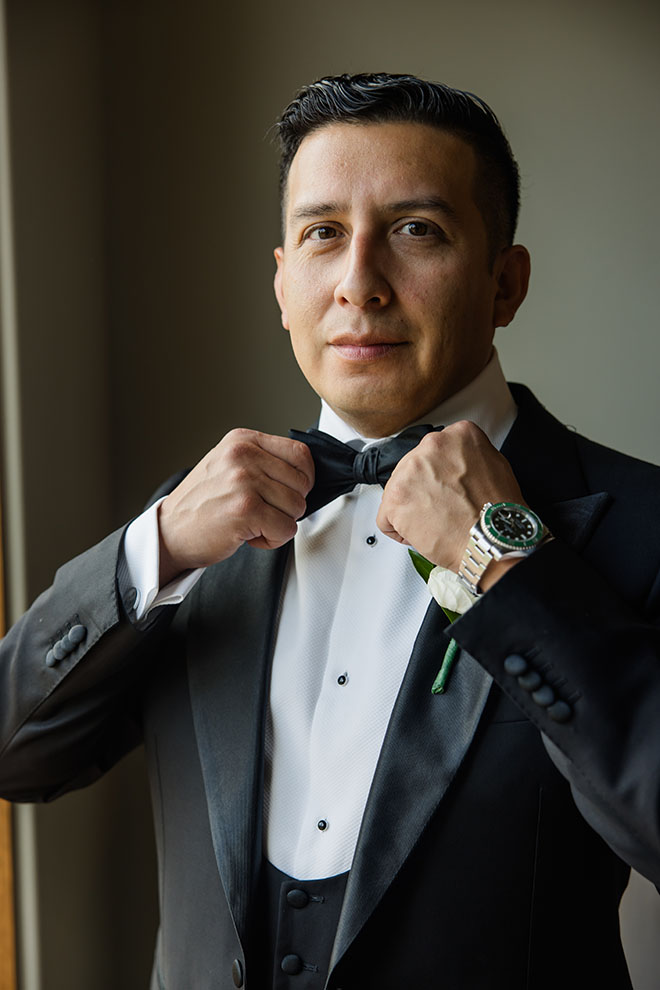 The groom adjusts his bow tie on his tuxedo. 