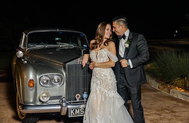 The bride and groom smile in front of a grey vintage car.