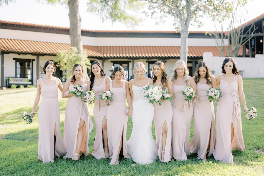 Bridesmaids pose for a group photo while the bride wears a white gown and the bridesmaids wear blush pink dresses. 