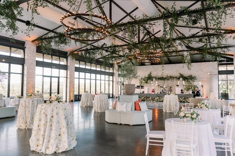 An inside wedding reception with high ceilings adorned in greenery, unique lighting, and colorful florals throughout the venue.