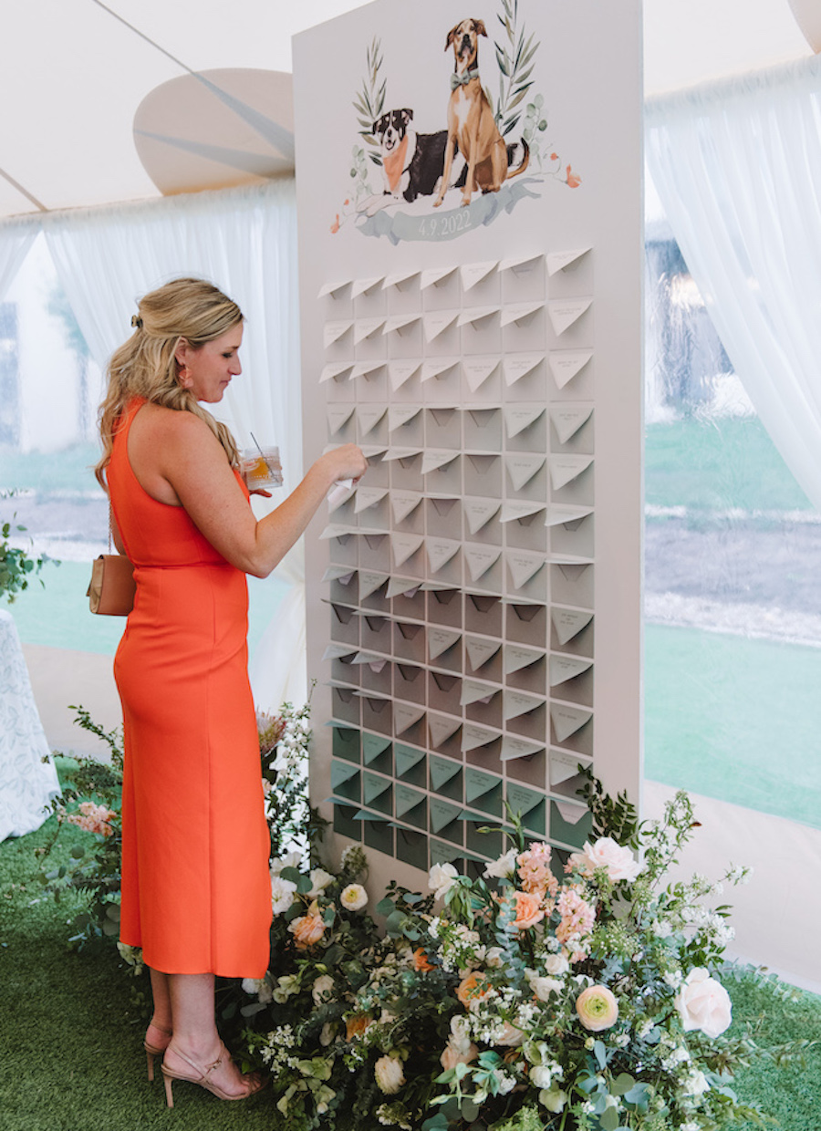 Guest stands near the guest table names at a wedding reception surrounded by florals and lush greenery. 