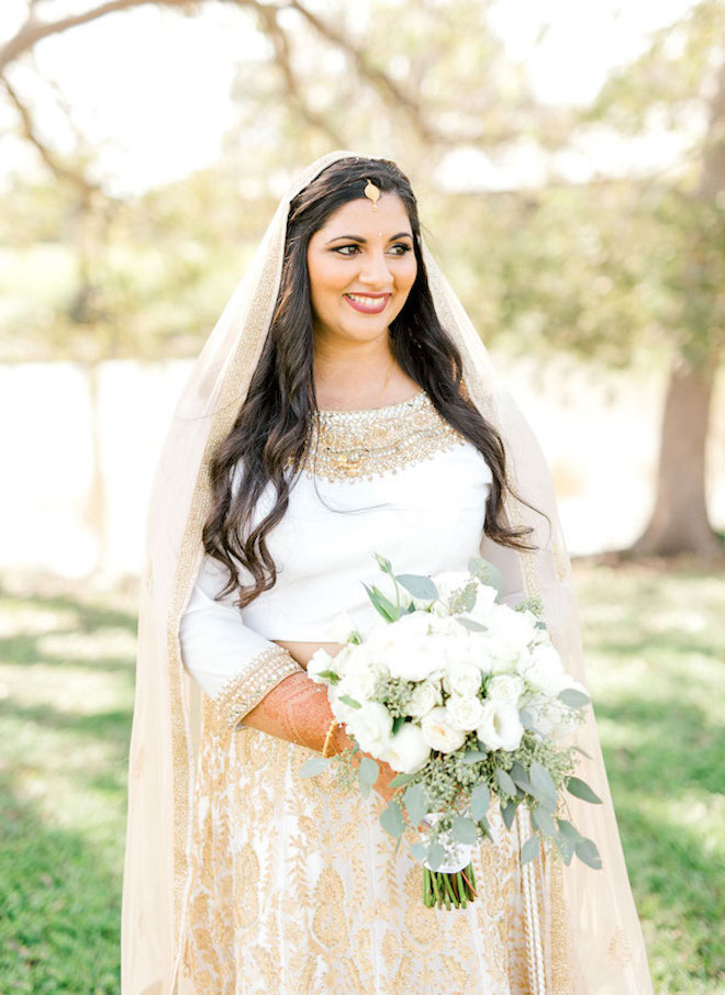 Bride dresses in a traditional Indian wedding gown, holding a white bouquet with a glam makeup look and skincare. 