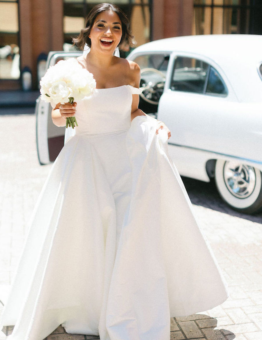 Bride smiles big for photo while she holds her gown and a white rose bouquet of flowers infront of a vintage car. 